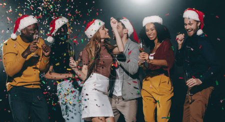 Téléchargez les photos : Heureux les jeunes dans des chapeaux de Noël profitant du champagne et dansant dans une boîte de nuit avec des confettis volant autour - en image libre de droit