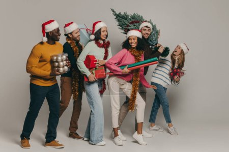 Foto de Jóvenes alegres llevando árbol de Navidad y varios ornamentos mientras caminan en el fondo del estudio - Imagen libre de derechos