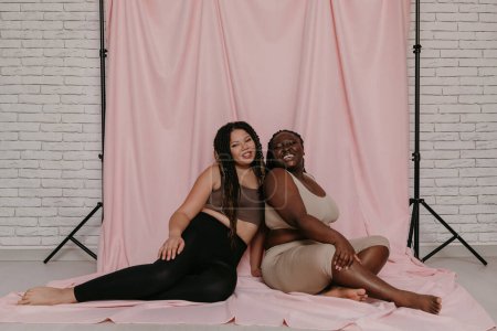 Foto de Dos mujeres africanas felices de talla grande en ropa deportiva sentadas y unidas sobre un fondo de tela rosa juntas - Imagen libre de derechos