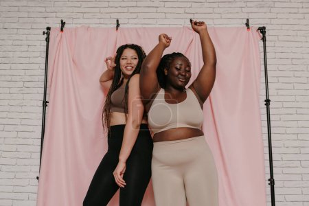 Téléchargez les photos : Deux femmes africaines souriantes de taille plus en tenue de sport dansant debout sur fond de tissu ensemble - en image libre de droit