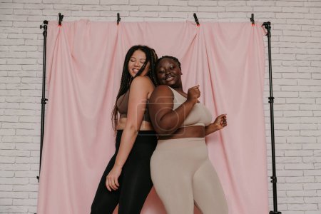 Foto de Dos sonrientes mujeres africanas de talla grande en ropa deportiva bailando mientras se paran espalda con espalda sobre un fondo de tela juntas - Imagen libre de derechos