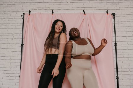 Téléchargez les photos : Deux femmes africaines heureuses de taille plus en tenue de sport dansant tout en se tenant sur fond de tissu ensemble - en image libre de droit