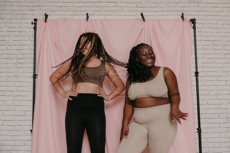 Photo for Two beautiful plus size African women in sportswear dancing while standing on fabric background together - Royalty Free Image
