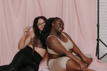 Téléchargez les photos : Deux femmes africaines ludiques plus la taille dans les vêtements de sport assis et collant sur fond de tissu rose ensemble - en image libre de droit