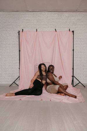 Téléchargez les photos : Deux femmes africaines heureuses de taille plus en vêtements de sport assis et collant sur fond de tissu rose ensemble - en image libre de droit