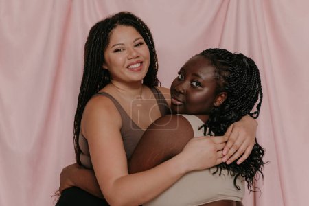 Téléchargez les photos : Deux belles femmes africaines de taille plus en vêtements de sport embrassant tout en se tenant sur fond de tissu - en image libre de droit