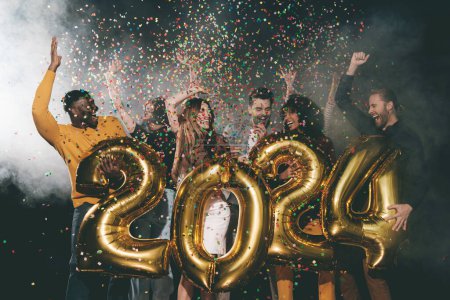 Foto de Grupo de jóvenes felices celebrando el año nuevo 2024 mientras lanzan confeti en el club nocturno - Imagen libre de derechos