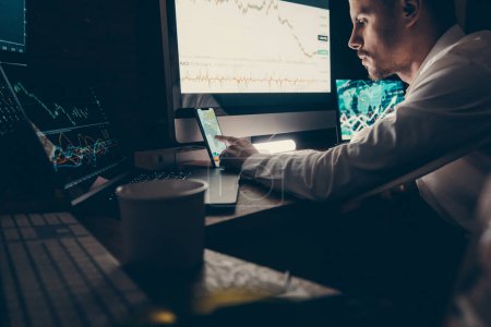 Foto de Comerciante del mercado de valores masculino utilizando tecnologías al analizar gráficos y gráficos en la oficina nocturna - Imagen libre de derechos