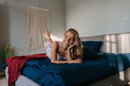Téléchargez les photos : Jeune femme détendue allongée au lit et souriante tout en passant du temps insouciant à la maison - en image libre de droit