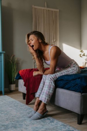 Photo for Beautiful young woman sitting on bed and yawning while waking up in the morning at home - Royalty Free Image