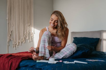 Téléchargez les photos : Joyeux jeune femme peignant des ongles de pieds tout en faisant pédicure sur le lit à la maison - en image libre de droit