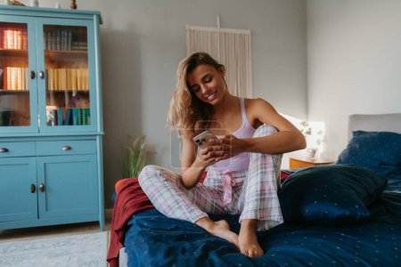 Photo for Relaxed young woman texting on smart phone and smiling while sitting on bed at home - Royalty Free Image