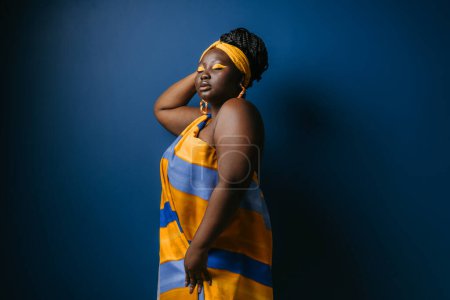 Téléchargez les photos : Belle femme africaine taille plus portant des vêtements africains traditionnels et des bijoux debout sur fond bleu - en image libre de droit