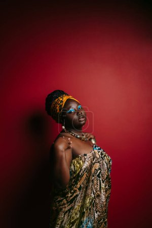 Téléchargez les photos : Superbe femme africaine taille plus avec un beau maquillage portant des vêtements traditionnels tout en se tenant debout sur fond rouge - en image libre de droit