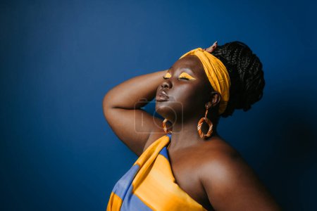 Photo for Attractive plus size African woman wearing traditional African attire and jewelry touching hair on blue background - Royalty Free Image