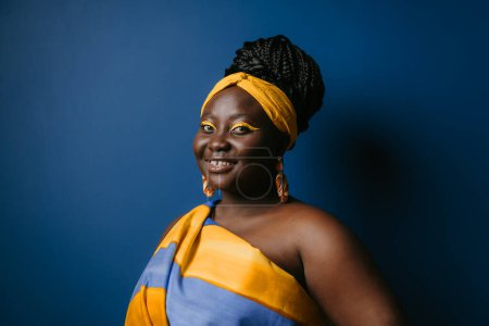 Téléchargez les photos : Souriant femme africaine taille plus portant des vêtements africains traditionnels et des bijoux en regardant la caméra sur fond bleu - en image libre de droit