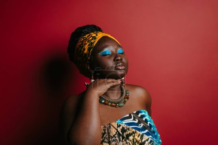 Téléchargez les photos : Superbe femme africaine taille plus avec un beau maquillage portant des vêtements traditionnels tout en se tenant debout sur fond rouge - en image libre de droit
