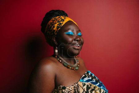 Foto de Preciosa mujer africana de talla grande con hermoso maquillaje usando ropa de cabeza tradicional sobre fondo rojo - Imagen libre de derechos