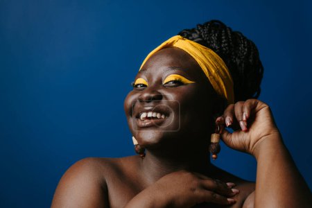 Téléchargez les photos : Portrait de sourire femme africaine taille plus avec belle boucle d'oreille de maquillage sur fond bleu - en image libre de droit
