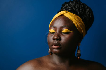 Téléchargez les photos : Portrait de magnifique femme africaine taille plus avec beau maquillage et bijoux debout sur fond bleu - en image libre de droit