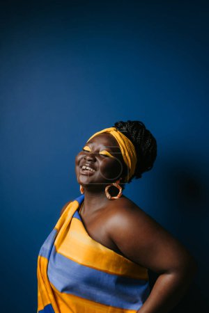 Téléchargez les photos : Confiante femme africaine taille plus portant des vêtements africains traditionnels et des bijoux souriant sur fond bleu - en image libre de droit