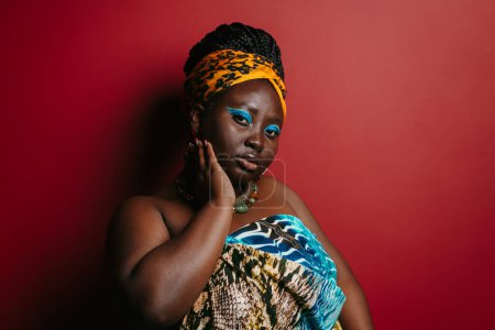 Téléchargez les photos : Superbe femme africaine taille plus avec un beau maquillage portant une tenue traditionnelle debout sur fond rouge - en image libre de droit