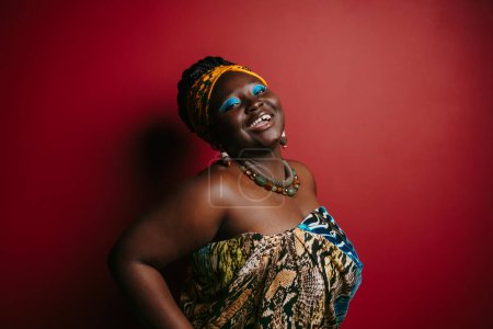 Foto de Feliz más tamaño mujer africana con hermoso maquillaje usando ropa de cabeza tradicional sobre fondo rojo - Imagen libre de derechos