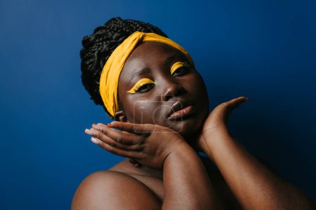 Téléchargez les photos : Portrait de femme africaine confiante avec beau maquillage et bijoux touchant visage sur fond bleu - en image libre de droit