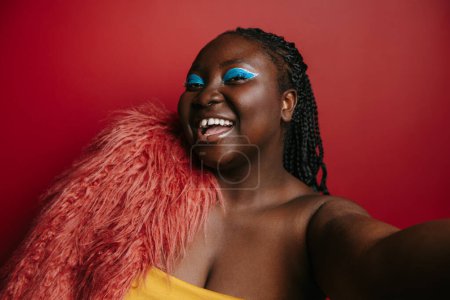 Foto de Moda más tamaño mujer africana con hermoso maquillaje sonriendo mientras se hace selfie sobre fondo rojo - Imagen libre de derechos