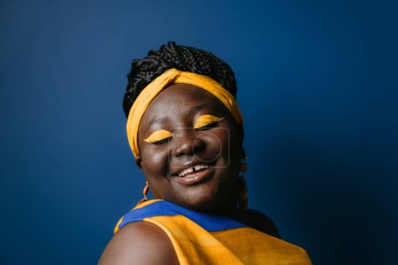 Téléchargez les photos : Femme africaine confiante de taille plus portant des vêtements et des coiffures traditionnels tout en souriant sur fond bleu - en image libre de droit