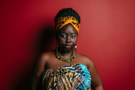 Téléchargez les photos : Confiante femme africaine taille plus portant des vêtements traditionnels et des bijoux en regardant la caméra sur fond rouge - en image libre de droit