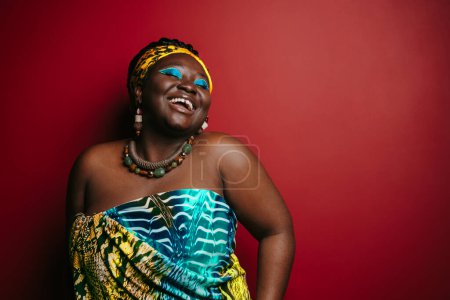 Téléchargez les photos : Souriant femme africaine taille plus avec beau maquillage portant des coiffures traditionnelles sur fond rouge - en image libre de droit