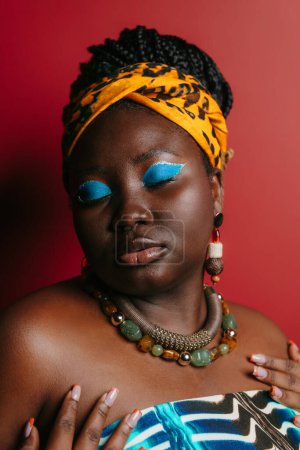 Foto de Moda más tamaño mujer africana con hermoso maquillaje usando atuendo tradicional sobre fondo rojo - Imagen libre de derechos