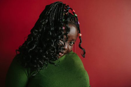 Téléchargez les photos : Femme africaine confiante avec des cheveux tressés et des clips colorés regardant la caméra sur fond rouge - en image libre de droit
