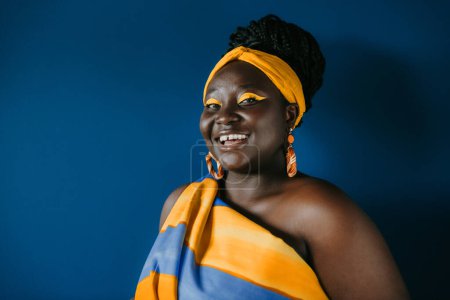 Téléchargez les photos : Souriant femme africaine taille plus portant des vêtements africains traditionnels et des bijoux en regardant la caméra sur fond bleu - en image libre de droit