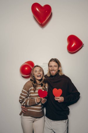 Photo for Smiling loving couple holding heart shape Valentines cards on beige background with balloons flying around - Royalty Free Image