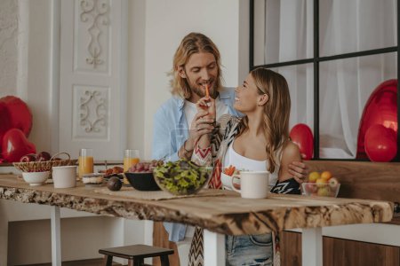 Foto de Mujer joven sonriente alimentando a su novio mientras ambos disfrutan del desayuno en casa decorado para el día de San Valentín - Imagen libre de derechos