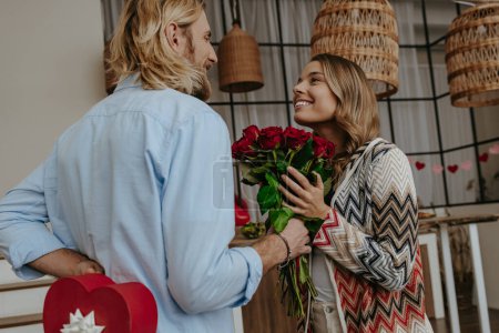 Foto de Joven hombre sonriente dando un ramo de rosas rojas a su novia sorprendida mientras esconde la caja de regalo a sus espaldas - Imagen libre de derechos