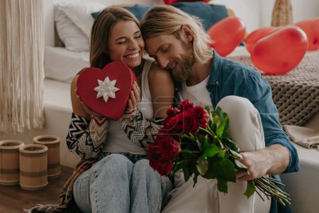 Foto de Amante pareja abrazando y sosteniendo rosas y caja de regalo mientras se apoya en la cama con globos en forma de corazón en el fondo - Imagen libre de derechos