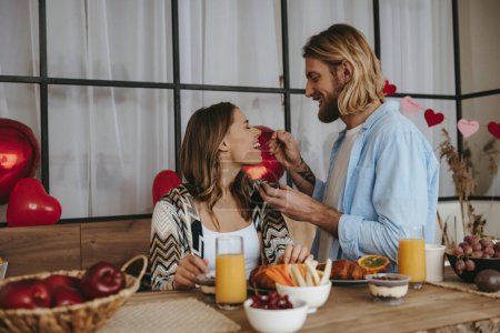 Foto de Sonriente joven alimentando a su novia con fruta de la pasión mientras disfrutan de un desayuno saludable en la cocina juntos - Imagen libre de derechos