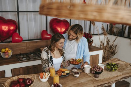 Téléchargez les photos : Vue du dessus du jeune couple souriant préparant le petit déjeuner à la cuisine avec des ballons rouges en forme de coeur sur le fond - en image libre de droit