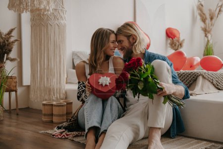 Photo for Loving couple embracing and holding roses and gift box while leaning on bed with heart shape balloons on background - Royalty Free Image