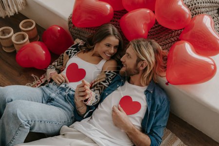 Photo for Top view of happy loving couple holding heart shape Valentines cards while leaning on bed surrounded with red balloons - Royalty Free Image