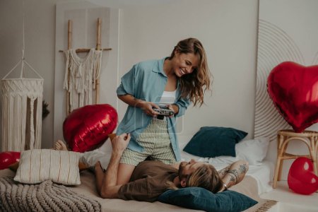 Téléchargez les photos : Belle jeune femme souriante photographiant son petit ami avec caméra rétro tout en se relaxant au lit à la maison - en image libre de droit