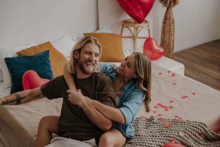 Téléchargez les photos : Couple aimant ludique embrasser et s'amuser tout en étant assis sur le lit entouré de ballons en forme de coeur rouge et confettis - en image libre de droit