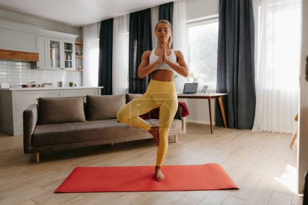 Téléchargez les photos : Pleine longueur de jeune femme confiante en vêtements de sport pratiquant le yoga tout en méditant à la maison - en image libre de droit