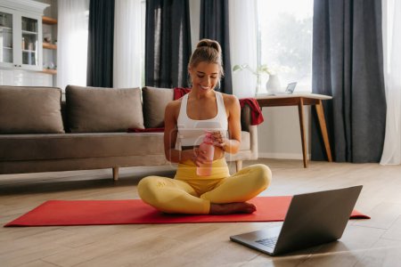 Téléchargez les photos : Femme sportive souriante tenant une bouteille d'eau tout en se reposant sur un tapis d'exercice à la maison avec un ordinateur portable au premier plan - en image libre de droit