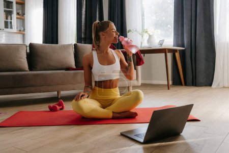 Téléchargez les photos : Heureuse femme sportive tenant une bouteille d'eau tout en se reposant sur un tapis d'exercice à la maison avec un ordinateur portable au premier plan - en image libre de droit