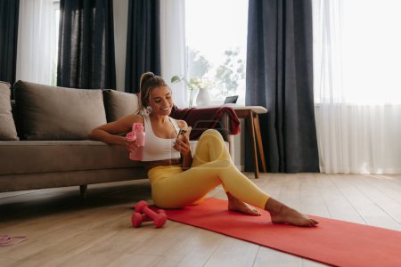 Photo for Happy sporty woman holding water bottle and using smart phone while resting on exercise mat at home - Royalty Free Image