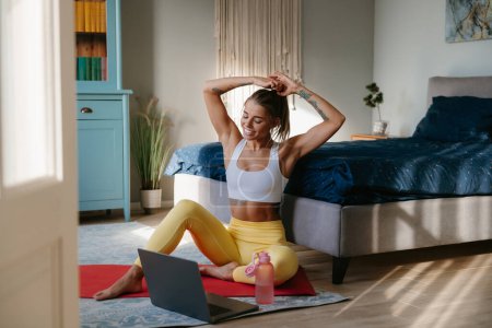 Téléchargez les photos : Heureuse femme sportive ajustant les cheveux et regardant ordinateur portable tout en se préparant à l'entraînement à la maison - en image libre de droit